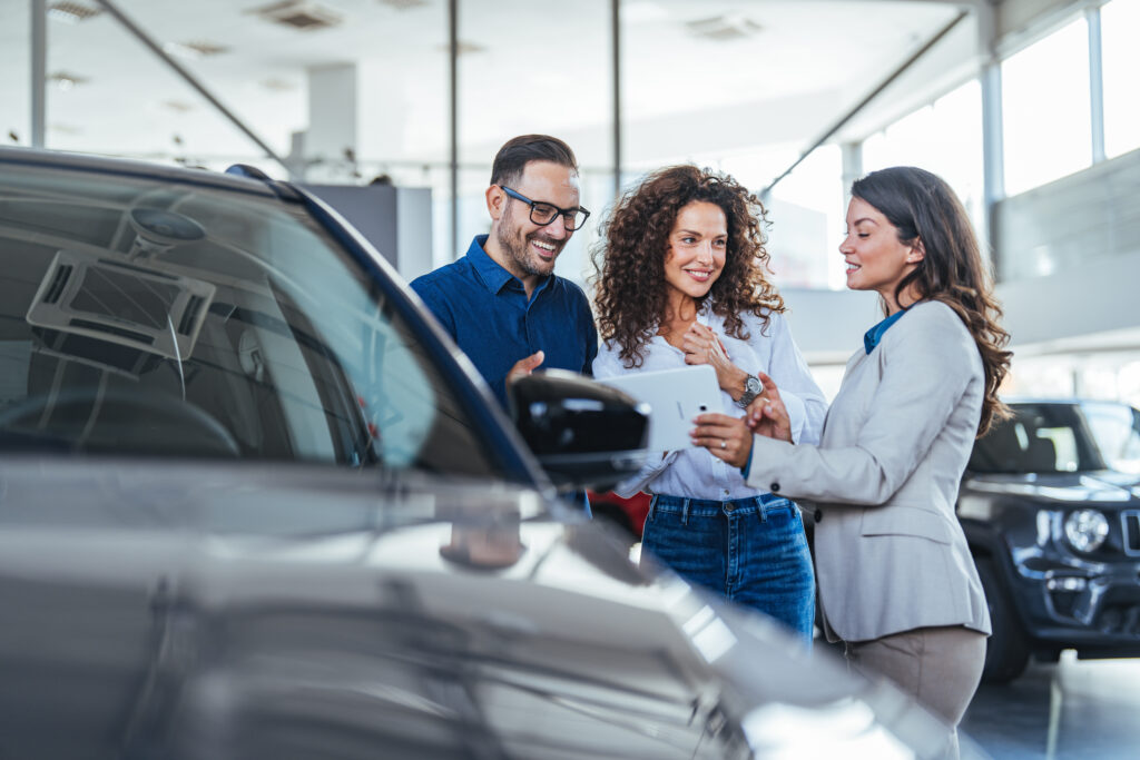 Friendly Group Of Friends In Car Showroom Review Tablet And Discuss Vehicle Purchase With Sales Team